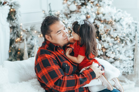 Asian family father dad hugging with daughter toddler girl celebrating Christmas or New Year. Mixed race dad man with kid sitting on couch sofa and cuddling at home. Winter holiday celebration.の写真素材