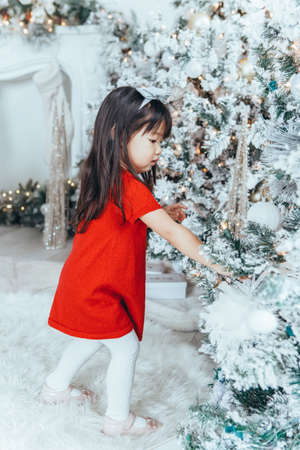 Asian toddler girl child in red dress decorating Christmas tree. Kid celebrating Christmas or New Year at home. Winter holiday celebration.の写真素材
