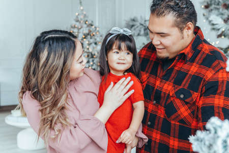 Asian family father, mother with daughter toddler girl celebrating Christmas or New Year. Mixed race mom, dad, daughter decorating Christas tree hugging at home. Winter holiday celebration.の写真素材