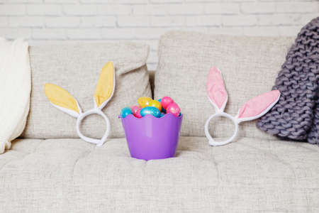 Basket with eggs and bunny ears headbands on couch sofa at home. Easter spring holiday celebrationの写真素材