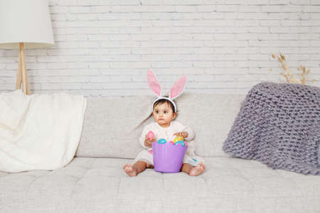 Cute Indian baby girl with pink bunny ears and basket of colorful eggs celebrating Easter holiday.の写真素材