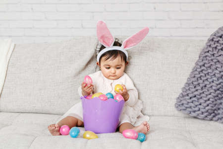 Cute Indian baby girl with pink bunny ears and basket of colorful eggs celebrating Easter holiday.の写真素材