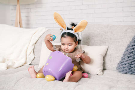 Cute Indian baby girl with pink bunny ears and basket of colorful eggs celebrating Easter holiday.
