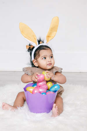 Cute Indian baby girl with pink bunny ears and basket of colorful eggs celebrating Easter holiday.の写真素材