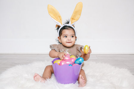 Cute Indian baby girl with pink bunny ears and basket of colorful eggs celebrating Easter holiday.の写真素材