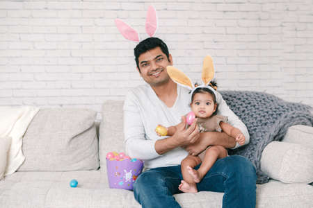 Indian father with baby girl daughter in bunny ears playing toy eggs in basket celebrating Easter.の写真素材