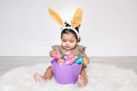 Cute Indian baby girl with pink bunny ears and basket of colorful eggs celebrating Easter holiday.の写真素材
