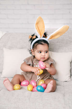 Cute Indian baby girl with pink bunny ears playing with colorful eggs candies toys celebrating Easter holiday.の写真素材