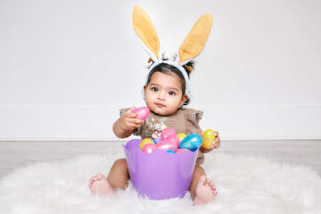 Cute Indian baby girl with pink bunny ears and basket of colorful eggs celebrating Easter holiday.の写真素材