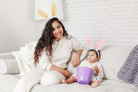 Indian mother with baby girl daughter in bunny ears playing toy eggs in basket celebrating Easter.の写真素材