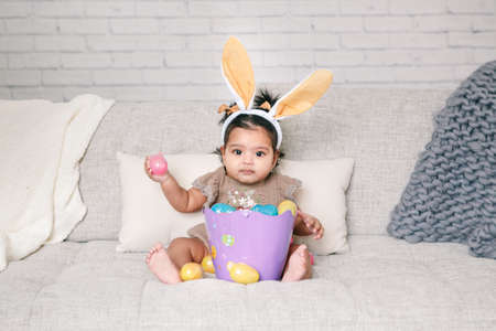 Cute Indian baby girl with pink bunny ears and basket of colorful eggs celebrating Easter holiday.の写真素材