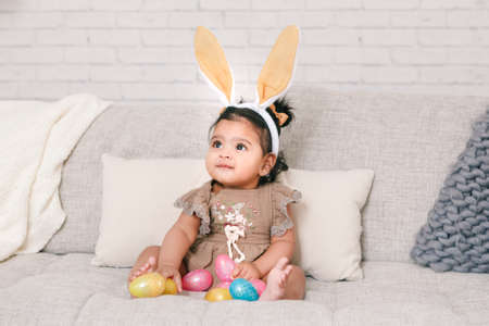 Cute Indian baby girl with pink bunny ears playing with colorful eggs candies toys celebrating Easter holiday.の写真素材
