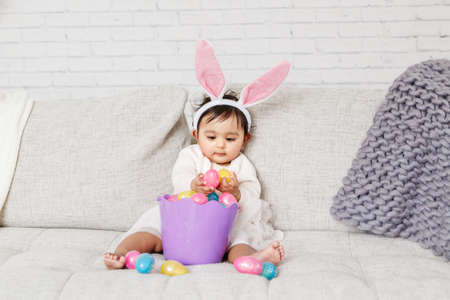 Cute Indian baby girl with pink bunny ears and basket of colorful eggs celebrating Easter holiday.の写真素材