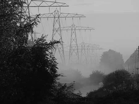 black and white photo of a foggy day, on the left are high-voltage pylons and on the right is a train track with associated poles, with trees in the foreground and a misty ditch in the middleの写真素材