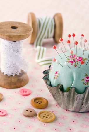 Green handmade floral pincushion in an old metal cupcake with buttons and spools of thread and lace, sewing concept backgroundの写真素材