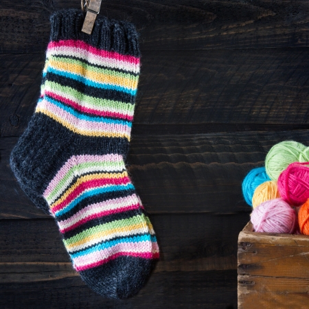 Pair of colorful striped woolen socks hanging on clothesline on dark wooden vintage background with multicolored balls of yarn and room for copy spaceの写真素材