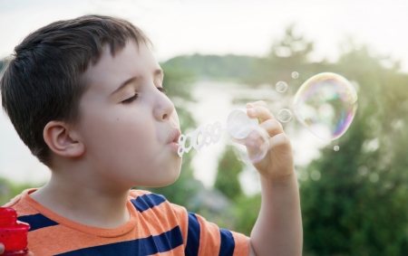Young boy blowing soap bubbles in summer evening sunlight with green nature background