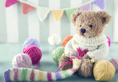 Teddy bear in a woolen sweater knitting a striped scarf with colorful balls of yarn