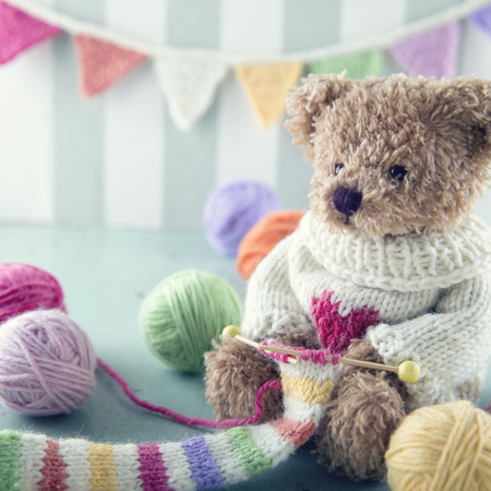 Teddy bear in a woolen sweater knitting a striped scarf with colorful balls of yarnの写真素材
