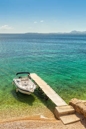 Empty boat standing at wooden pier under bright sunlight with shadow on pebbles at sea floor seen through transparent water of Ionian sea, Corfu, Greeceの写真素材
