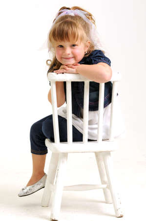 young pretty blond girl with blue dress sitting on a white chair and smilingの写真素材