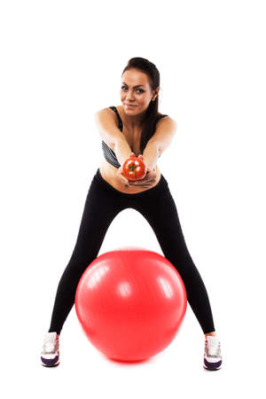 Slim girl wearing training suit sitting on red gym ball offering a tomato, isolated on white, healthy life-style promotionの写真素材