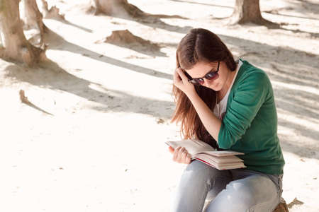 Beautiful young brunette woman wearing green sweater and sun glasses is carried away by reading a book in the parkの写真素材