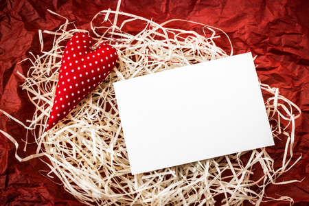 Blank white paper card for copy space and vintage handmade red valentines day toy heart over pile of straw and crumpled paper backgroundの写真素材