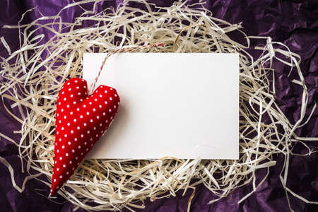 Blank white paper card for copy space and vintage handmade red valentines day toy heart over pile of straw and crumpled paper backgroundの写真素材