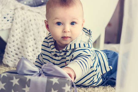 Portrait of a cute baby boy with big blue eyes wearing jeans and striped hoodie sweater and reaching to his present in wrapped box with ribbon. Birthday or Christmas presents conceptの写真素材