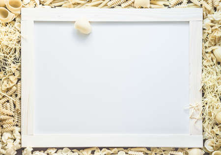 Mixed dried pasta selection surrounding white board with wooden frame for your text. Top view shot