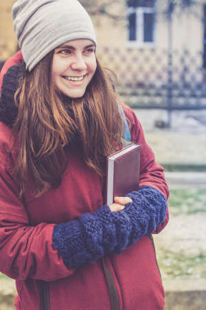 Beautiful hipster girl with long brown hair, holding book in her hands in winter clothes outdoor...