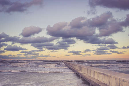 Perspective view of a concrete pier at the sea at sunset with dramatic sky and rolling waves. Stormy weather, forces of nature conceptの写真素材