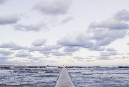 Perspective view of a concrete pier at the sea at sunset with dramatic sky and rolling waves. Stormy weather, forces of nature conceptの写真素材