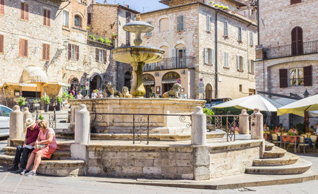 ASSISI, ITALY - SEPTEMBER 4, 2016: Couple of tourists sitting at the historic fountain figuring three lions and medieval buildings in the background on Piazza del Comune the main square of Assisi, Umbria, Italyのeditorial素材