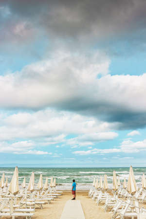 Lonely man is standing on the empty beach among the rows of closed umbrellas and deckchairs. The beginning or back-end of the season concept.の写真素材