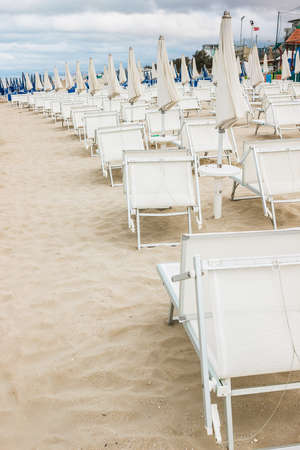 Rows of closed white umbrellas and deckchairs on the empty beach before the storm. The beginning or back-end of the season concept.の写真素材