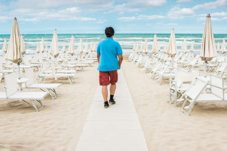 Lonely man walking on the empty beach among the rows of closed umbrellas and deckchairs. The beginning or back-end of the season concept.の写真素材