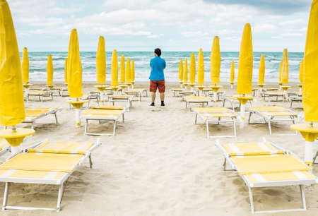 Lonely man is standing on the empty beach among the rows of closed umbrellas and deckchairs. The beginning or back-end of the season concept.の写真素材