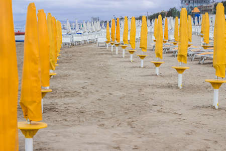 Rows of closed orange umbrellas and deckchairs on the empty beach before the storm. The beginning or back-end of the season concept.の写真素材