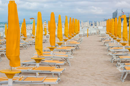 Rows of closed orange umbrellas and deckchairs on the empty beach before the storm. The beginning or back-end of the season concept.の写真素材