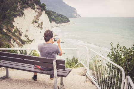 Behind view of young man using smart phone for taking photo of seaside panorama in Numana, Marche, Italy. Travel conceptの写真素材