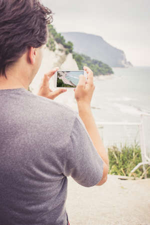 Young man using smart phone for taking photo of seaside panorama in Numana, Marche, Italy. Travel conceptの写真素材