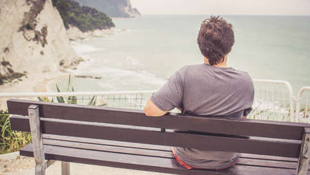 Back side view of alone young man sitting on bench overlooking sea and Mount Conero, Numana, Marche, Italy.の写真素材
