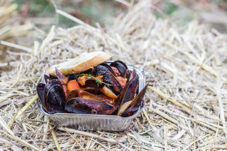 Prepared mussels served with vegetables and a slice of bread in foil box on a stack of strawの写真素材