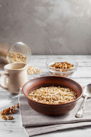 Oat flakes in brown clay bowl ready to cook, scattered oat flakes, walnuts, jug with milk and spoon on the light wooden tableの写真素材