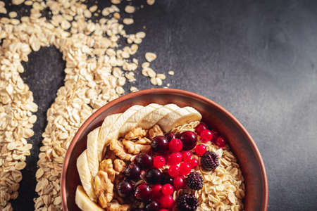 Oatmeal in brown clay bowl with berries (blackcurrants, redcurrants, blackberries), bananas and walnuts ready to eat, silhouette of spoon on scattered flakes over black backgroundの写真素材