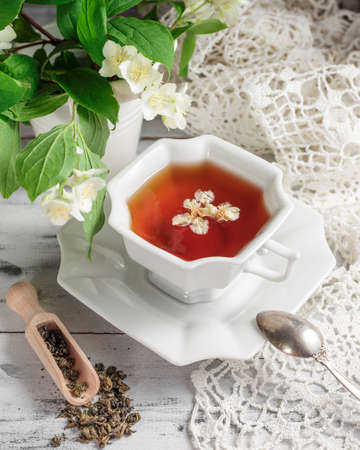 White porcelain cup of tea with jasmine flowers, scattered dry tea leaves and wooden spoon on a white wooden table. Selective focus on top of the cupの写真素材