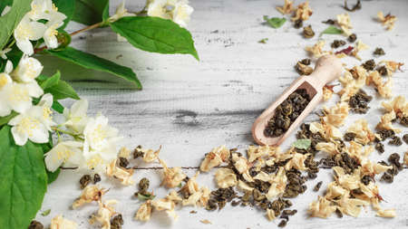 Scattered dried green tea leaves with fresh jasmine flowers and wooden spoon on a white wooden tableの写真素材