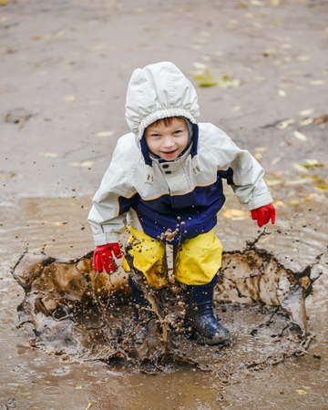 Happy two years old boy jumping on muddy puddles in rubber boots